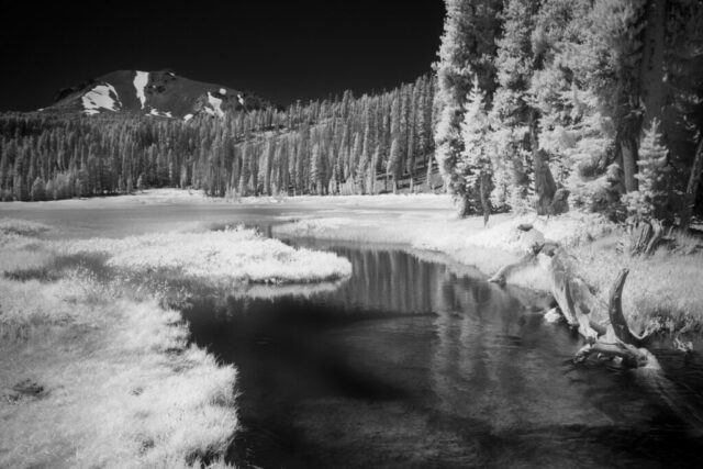 Black and white deep infrared photograph of Kings Creek winding through a mountain meadow beneath Lassen Peak in Lassen Volcanic National Park, California. Captured with an 830nm deep infrared filter on a full spectrum modified camera, the image reveals luminous white foliage and reflective water beneath a dark, dramatic sky. The snow patches on the volcanic slopes and glowing pines create a surreal, high-contrast alpine landscape characteristic of fine art infrared photography.