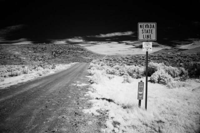 Black and white deep infrared photograph of a dirt road leading to the Nevada state line near Smoke Creek Road, on the border of California and Nevada. Captured with an 830nm infrared filter on a full spectrum modified camera, the image shows glowing desert brush beneath a dark, high-contrast sky. The bold “Nevada State Line” sign stands prominently in the foreground, creating a stark and timeless Western landscape scene.