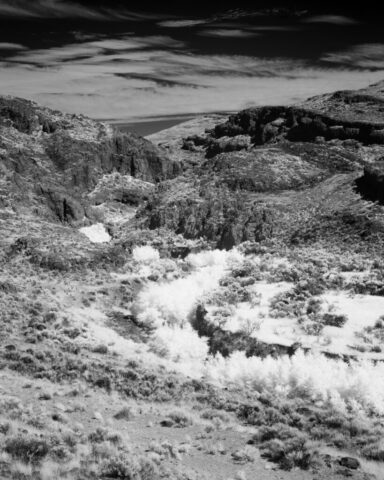 Black and white deep infrared landscape photograph of Rush Creek winding through the basalt canyon of the Smoke Creek Desert in Washoe County, Nevada, near the California border. Captured with an 830nm infrared filter on a full spectrum modified camera, the image features glowing white creekside foliage set against rugged desert terrain. The high-contrast rendering emphasizes texture, light, and the stark beauty of this historic region along the Nobles Immigrant Trail.