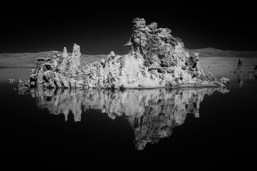 Black and white deep infrared photograph of tufa limestone formations reflected in the calm waters of Mono Lake, California. Captured with an 830nm “deep infrared” filter on a full spectrum modified camera, the image reveals dramatic contrast and intricate texture under bright midday light. The luminous tufa spires and darkened sky evoke the timeless mood of classic monochrome landscape photography, reminiscent of Ansel Adams’ high-contrast style.