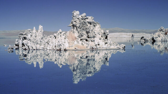 The Invisible Light of Noon Infrared landscape photograph of tufa formations at South Tufa on Mono Lake, California, captured in late morning light with a 590nm near-infrared filter on a full spectrum modified camera. The surreal image shows pale limestone spires reflected in calm blue water beneath a clear summer sky. The infrared spectrum reveals striking tonal contrast and clarity, transforming this otherworldly scene into an ethereal fine art composition.