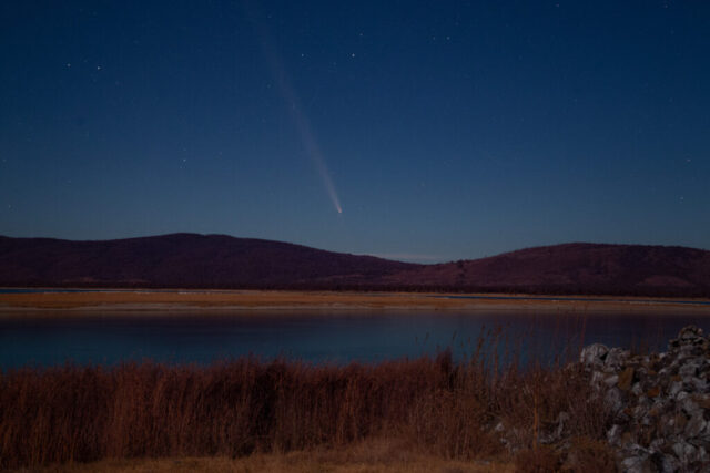 Cosmic Lake Night landscape photograph of comet C/2023 A3 Tsuchinshan-ATLAS glowing above Eagle Lake in Lassen County, California. Captured on a clear October 2024 evening, the long-exposure image shows the comet’s bright nucleus and glowing tail streaking upward in the deep blue night sky. The lake reflects the moonlit colors of the surrounding hills and the golden marsh grasses in the foreground, with remnants of old dredger piles adding texture and historical depth. Illuminated by a nearly full moon, this astrophotography scene blends celestial motion and quiet terrestrial beauty near the Chico State Biological Camp at Eagle Lake, highlighting the contrast between cosmic wonder and the serene Northern California landscape.