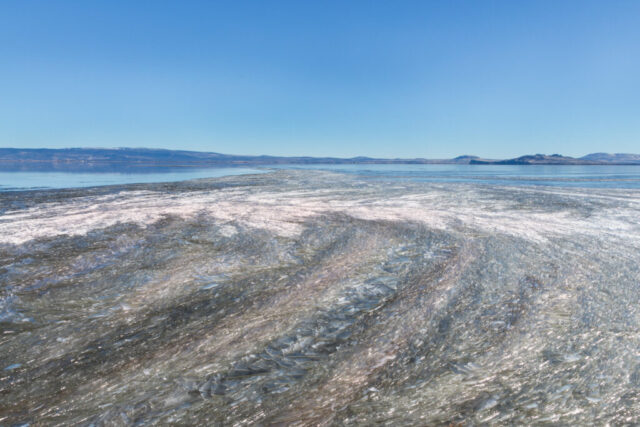 Winter landscape photograph of Tule Lake National Wildlife Refuge in Northern California, showcasing intricate patterns of wind-sculpted ice formations near the edge of the water. Captured on a mild January day along the refuge’s scenic auto tour route, the image reveals layers of crystalline textures shimmering under clear blue skies, framed by the distant ridges of the Klamath Basin. The frozen surface tells a story of shifting winds and changing light, creating a mesmerizing natural mosaic. A tranquil study of form, texture, and reflection—celebrating the quiet, elemental beauty of Tule Lake in winter.
