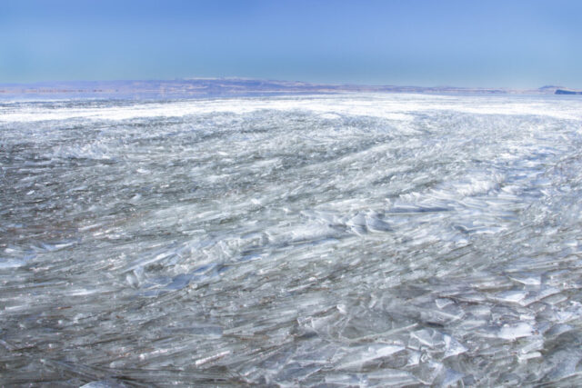 Winter landscape photograph of Tule Lake National Wildlife Refuge in Northern California, featuring a mesmerizing expanse of layered ice formations stretching across the frozen surface. Captured on a calm January day under brilliant blue skies, the scene reveals the natural artistry created by shifting winds and fluctuating temperatures—each shard of ice glinting in the sunlight like cut glass. The distant ridges of the Klamath Basin form a subtle backdrop, grounding this abstract pattern of nature’s geometry. A serene and captivating study of texture, reflection, and the quiet stillness of Tule Lake in midwinter.
