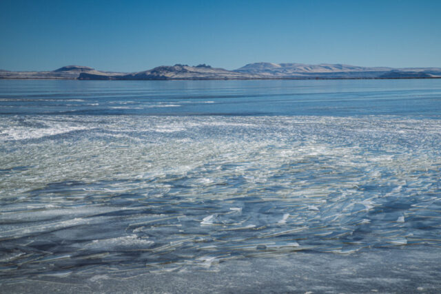 Landscape photograph of Tule Lake National Wildlife Refuge in Northern California, depicting intricate sheets of wind-sculpted ice shimmering beneath a clear blue winter sky. Captured on a calm January afternoon, the fractured ice catches and reflects sunlight like scattered glass, leading the eye toward the distant snow-dusted ridges of the Klamath Basin. The tranquil stillness of the frozen lake contrasts beautifully with the subtle sense of movement left behind by shifting winds. A serene, textural study of Tule Lake’s winter beauty—quiet, vast, and ever-changing.