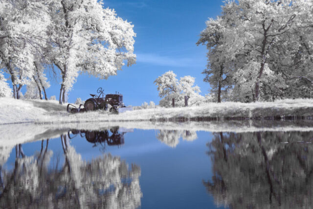 Rural Visions in Invisible Light False color infrared photograph of a vintage tractor beside a reflective pond on a Shasta County ranch, captured with a 720nm infrared filter on a full spectrum modified camera. The near-infrared light turns blue oak foliage nearly white under a deep blue sky, creating a surreal, dreamlike rural landscape mirrored perfectly in still water.