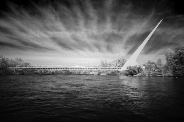 Sundial Bridge in Invisible Light Infrared black and white photograph of the Sundial Bridge in Redding, California, captured with an 830nm deep infrared filter on a full spectrum modified camera. The bridge’s dramatic cable-stay pylon rises above the Sacramento River beneath streaking high clouds rendered dark against bright white foliage, creating a striking, high-contrast composition blending architecture and nature.