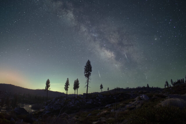 Lyrids and Milky Way over Plumas County Astrophotography image of the Lyrid meteor shower and the Milky Way rising over Plumas County, California, captured from the Janesville Grade near Antelope Lake. Two bright meteors streak through the dense star field of the galactic core, framed by tall pines and rugged terrain under a soft pre-dawn glow. The faint light along the horizon is a waning crescent moon, illuminating the rocky landscape without overpowering the stars. This serene nightscape blends cosmic motion and earthly stillness, highlighting the natural beauty of Northern California’s mountain skies during the April Lyrids meteor shower, one of the oldest recorded celestial events in human history.