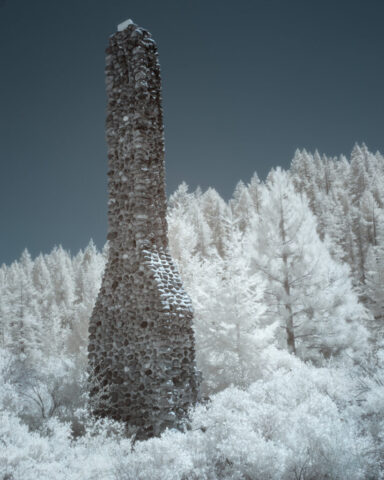 Infrared photograph of an old stone chimney along California State Route 49 near Sierra City, captured with a 720nm near-infrared filter. The weathered chimney rises among bright white trees and foliage, creating a surreal and haunting Sierra landscape.