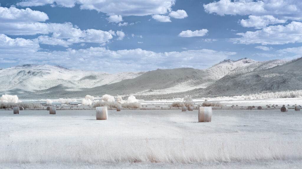 Infrared photograph of a hay field with large round bales under a sky of dramatic clouds and distant Sierra Nevada foothills. Captured with a 720nm near-infrared filter, the landscape glows with soft, surreal tones of white foliage and pale blue sky.