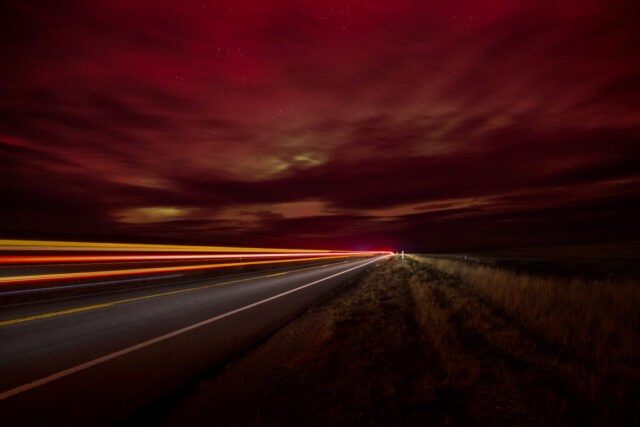 On a Dark Desert Highay... Long-exposure night photograph along a desolate stretch of US Highway 395 under a dark red sky faintly illuminated by aurora activity. The road disappears into the horizon beneath a glowing canopy of clouds tinged with pink and green hues. Streaks of red, yellow, and white light trails from a passing semi-truck cut across the frame, leading the viewer’s eye toward the distant vanishing point. Dry desert grass lines the roadside, and faint stars scatter across the sky, creating an atmospheric, moody scene that captures the quiet beauty of a late-night drive through the high desert during an unexpected aurora display.