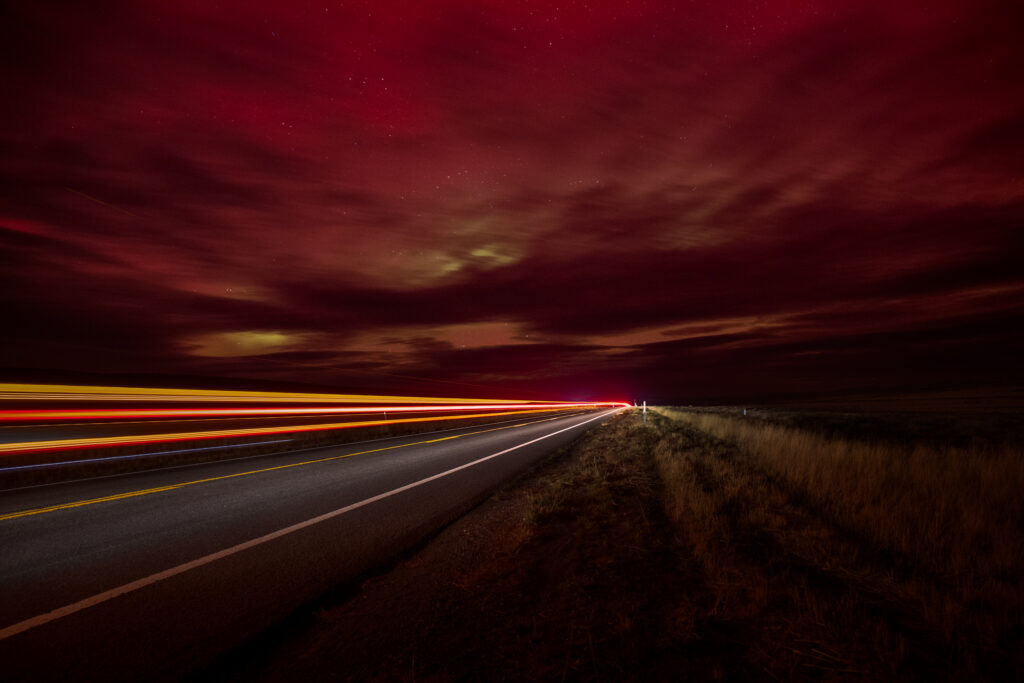 Long-exposure night photograph along a desolate stretch of US Highway 395 under a dark red sky faintly illuminated by aurora activity. The road disappears into the horizon beneath a glowing canopy of clouds tinged with pink and green hues. Streaks of red, yellow, and white light trails from a passing semi-truck cut across the frame, leading the viewer’s eye toward the distant vanishing point. Dry desert grass lines the roadside, and faint stars scatter across the sky, creating an atmospheric, moody scene that captures the quiet beauty of a late-night drive through the high desert during an unexpected aurora display.