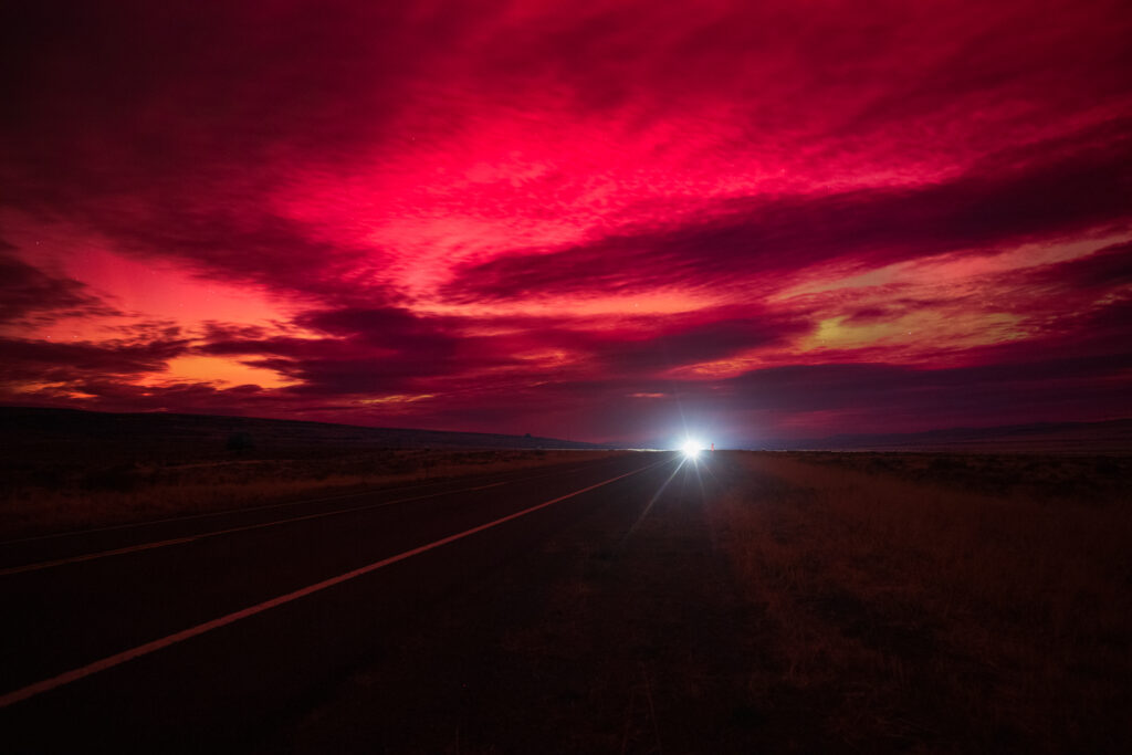 A long exposure photograph of US Highway 395 in Lassen County, California, taken during the November 11, 2025 geomagnetic storm. The image shows a glowing red and magenta sky from aurora reflections and solar flare activity, spreading across layered clouds above an open desert landscape. A single pair of headlights in the distance creates a bright focal point on the dark asphalt, with faint illumination revealing roadside grass and the vanishing lines of the highway. The dramatic, fiery sky dominates the scene, blending atmospheric light with the quiet isolation of the high desert night.