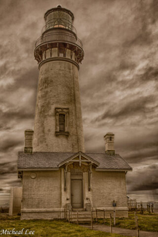 Yaquina Head Lighthouse rises above its keeper’s house on the Oregon coast near Newport, framed by dramatic overcast skies, weathered stonework, and a grassy foreground, with the tall cylindrical tower dominating the vertical composition.