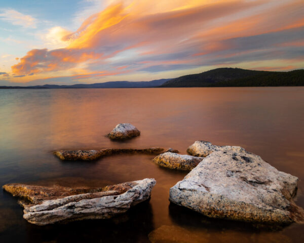 Moody Pikes Point Evening Flat volcanic rocks rest in shallow water along the shoreline at Pike’s Point on Eagle Lake in Lassen County, California, with smooth lake surface, distant forested hills, and streaked clouds glowing orange and pink at sunset.