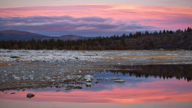 Pastel Reflections at Eagle Lake Pastel pink and purple sunset clouds reflected in shallow pools on a rocky shoreline near Eagle Lake in Lassen County, California, with forested hills and distant ridges along the horizon.