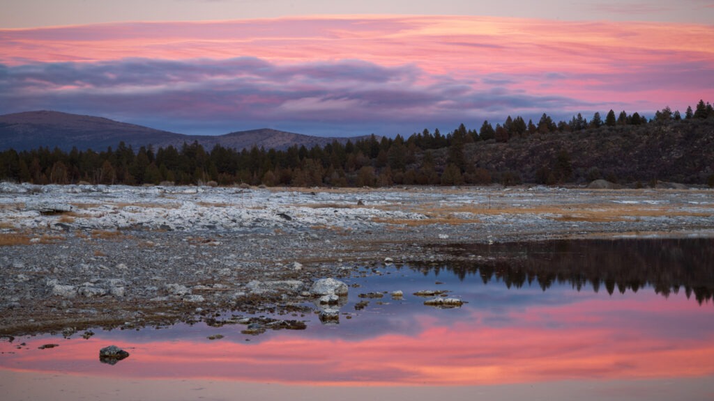 Pastel pink and purple sunset clouds reflected in shallow pools on a rocky shoreline near Eagle Lake in Lassen County, California, with forested hills and distant ridges along the horizon.