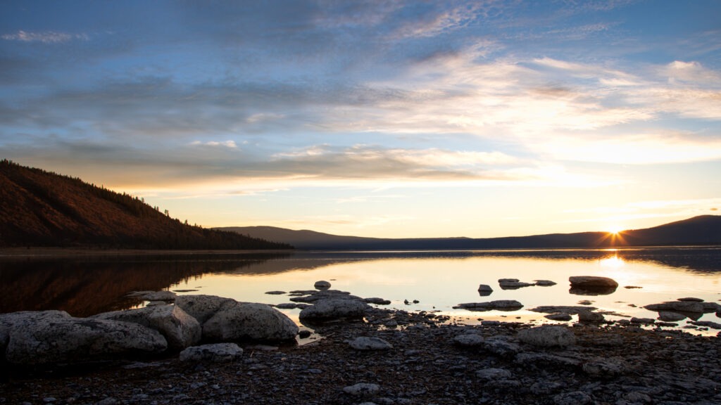 Sunset at Miner’s Point on the eastern shore of Eagle Lake in Lassen County, California, showing warm light across the western ridge, calm reflective water, and scattered pale rocks along the shallow shoreline.