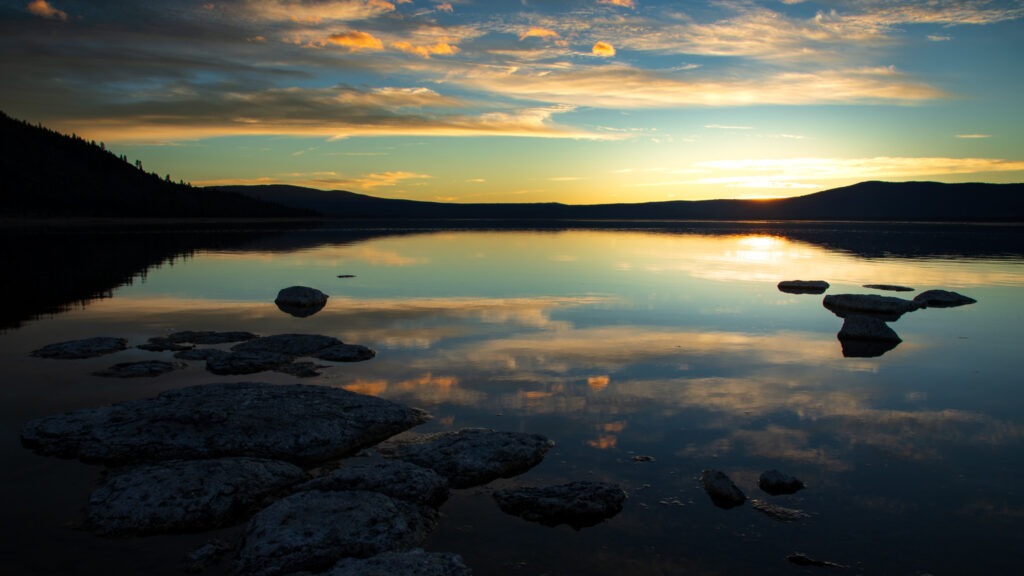 Sunset reflections on the calm, shallow water at Miner’s Point on the eastern shore of Eagle Lake in Lassen County, California, with scattered shoreline rocks and the sun dipping behind the western ridge.