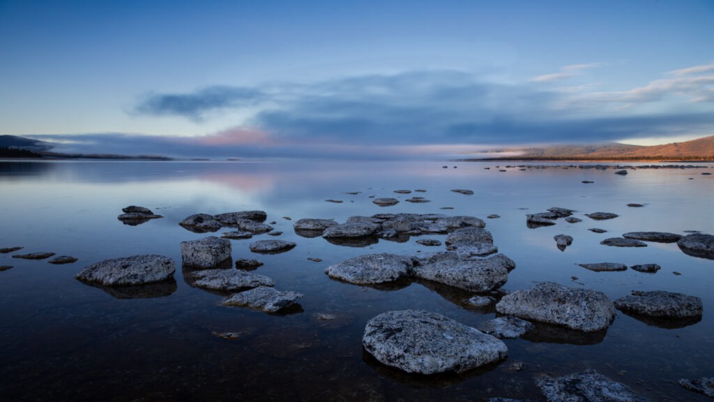 Calm winter morning at Eagle Lake near Miner’s Point on the east shore in Lassen County, California, with smooth water reflecting pastel dawn colors, distant fog, and scattered rocks in the shallow foreground under a clear blue sky.