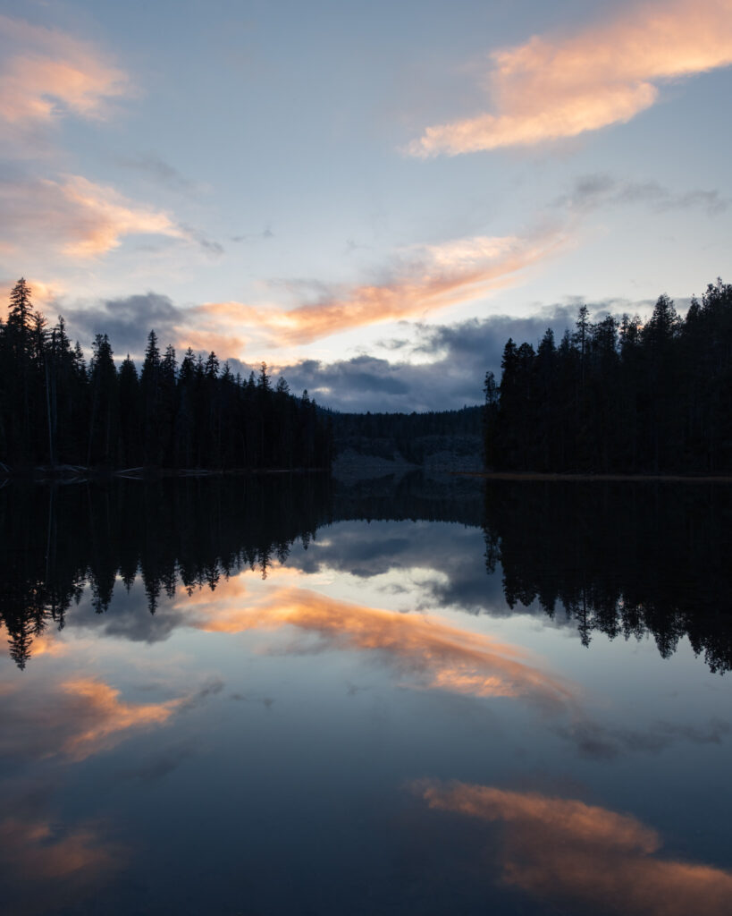 Symmetrical sunset reflections on calm Silver Lake in Lassen County, California, with dark pine forest lining both shores, low clouds on the horizon, and soft orange and pink clouds mirrored in the still water beneath a pale blue evening sky.
