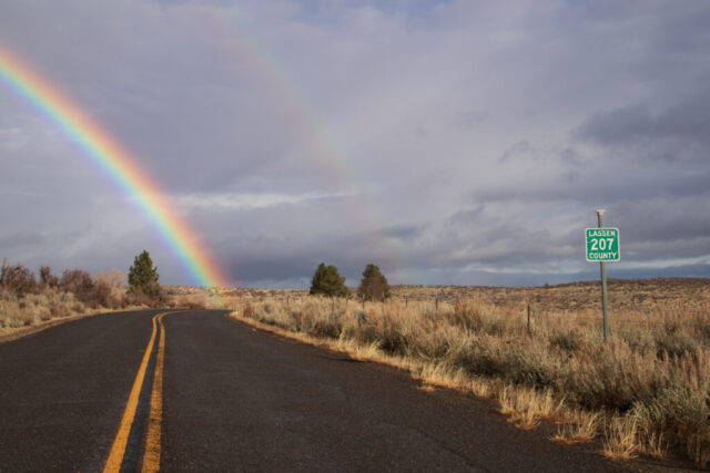 Rainbow on a Country Road - Lassen County California A vivid rainbow arcs across a cloudy sky above an empty rural road, with a green Lassen County 207 road sign on the right and dry high desert vegetation lining both sides of the pavement near Susanville, California.