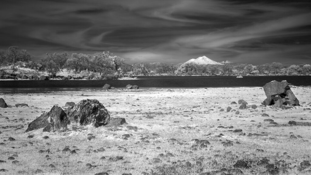 While driving along State Route 36 a few miles east of Red Bluff, California, at around 1pm on a sunny February day, I noticed the conditions looked very nice for infrared photography. The clouds were streaky and wispy and the sun was bright but somewhat filtered by the high haze. As I drove by the Sacramento Riverbend Area I could see the snow-covered Mount Shasta in the distance beyond Hog Lake. That clinched it, and I pulled in to stretch my legs, break out my full spectrum gear, and try for a shot or two. After capturing a wider view looking across the Sacramento Riverbend Area toward Mount Shasta, I moved closer to the shore of Hog Lake and recomposed around one of the larger volcanic boulders that litter the foreground. Shasta is still on the small side in the frame here, but a bit more prominent above the lake — its snow-covered summit catching the light while the dark infrared water creates a strong horizontal divide between the two halves of the image. The 830 nanometer filter renders water almost completely black due to its near-total absorption of infrared light, which gives the lake a graphic, almost inky quality that I find really satisfying in compositions like this. The foreground rock, by contrast, picks up texture and detail beautifully at this wavelength. Ansel Adams comes to mind — that kind of extreme tonal separation between a dark sky, bright foliage, and a strong foreground element is very much in his wheelhouse, even if he got there with a deep red filter and Kodak rather than a modified Canon 6D. The Sacramento Riverbend Area is BLM-managed land with documented human presence going back 6,000 to 10,000 years, sitting at the old boundary between the Yana and Nomlaki peoples. It is a genuinely underrated spot.